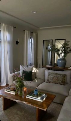 a living room with white couches and pillows on top of the coffee table in front of two windows