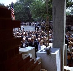 a large group of people standing in front of a building with an american flag hanging from it's side