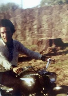 a man riding on the back of a motorcycle down a dirt road next to a hillside