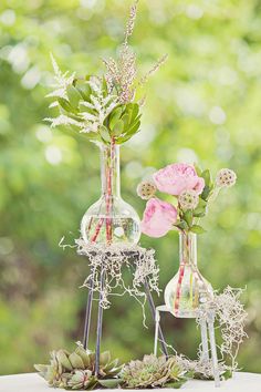 two vases filled with flowers and plants on top of a white cloth covered table