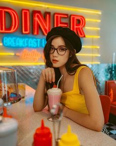 a woman sitting at a table with a drink in front of her and a neon sign behind her