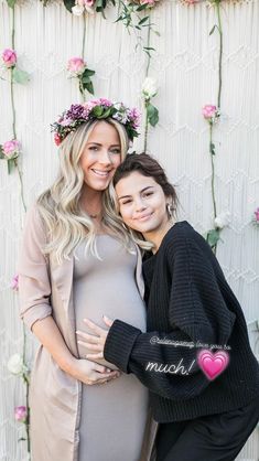 two pregnant women pose for the camera in front of a wall with flowers and greenery