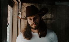 a man with long hair wearing a cowboy hat and white t - shirt standing in front of a wooden wall