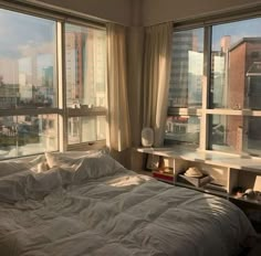 an unmade bed in front of large windows with cityscape outside the window