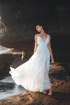 a woman in a white dress standing on rocks near the water and looking off into the distance