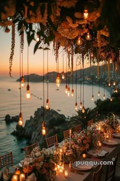 an outdoor dinner table with candles and flowers hanging from it's ceiling over the ocean