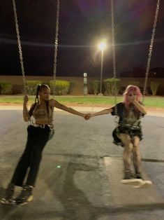 two young women holding hands while riding skateboards on a swing at night in a parking lot