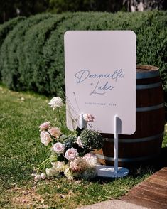 a sign sitting on the ground next to a barrel with flowers and greenery around it