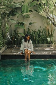 a woman sitting on the edge of a pool in front of some plants and trees