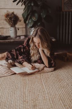 a woman laying on top of a bed next to a plant and writing in a book