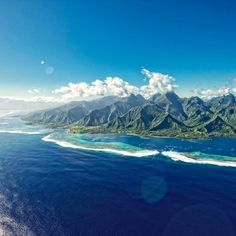 an aerial view of the ocean with mountains in the background and blue skies above it