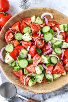 a wooden bowl filled with cucumber, tomatoes and onions next to two spoons