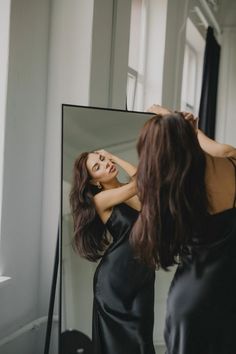 a woman standing in front of a mirror looking at her reflection wearing a black dress