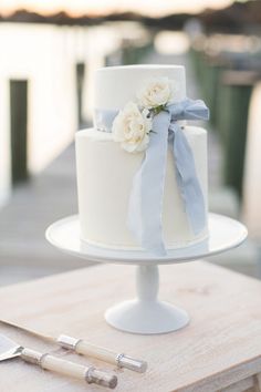 a white wedding cake with blue ribbon and flowers on a wooden table next to the water