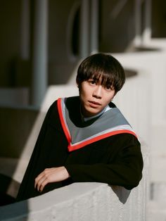 a young man leaning against a white wall wearing a black and grey graduation gown with red trim