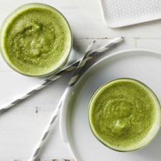 two glasses filled with green smoothie sitting on top of a white plate next to straws