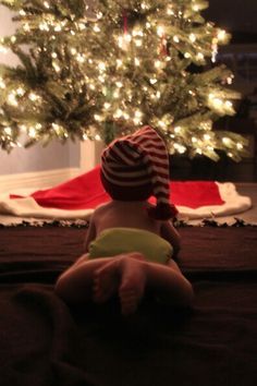 a baby sitting on the floor in front of a christmas tree wearing a striped hat