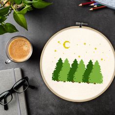 a cross stitch pattern with green trees and the moon in the sky next to a cup of coffee