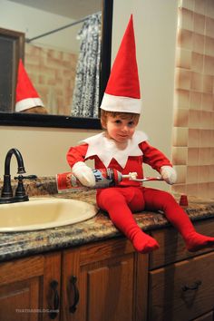 a young child sitting on top of a bathroom sink wearing an elf hat and holding a toothbrush