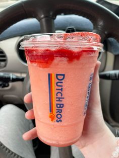 a person holding up a pink drink in their hand and steering wheel behind the car