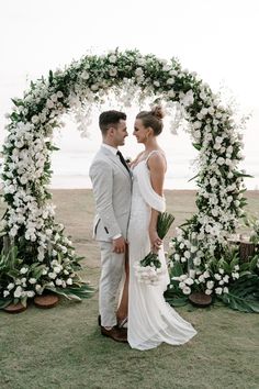 a man and woman standing in front of a wedding arch with white flowers on it