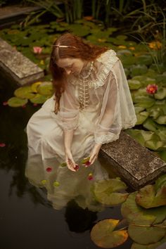 a woman is sitting in the water surrounded by lily pads and pink flowers, looking down at her hands