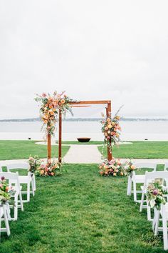 an outdoor ceremony set up with white chairs and floral arrangements on the aisle, overlooking water