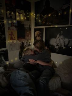 two people sitting on a bed hugging each other with lights strung all over the wall behind them