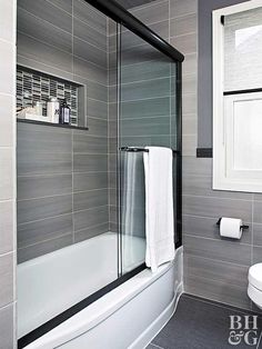 a white toilet sitting next to a bath tub under a bathroom window in a gray tiled bathroom