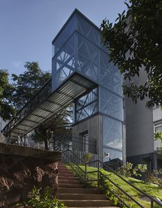 the stairs lead up to the top of this modern glass - walled building, which is surrounded by greenery and trees