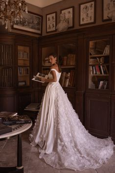 a woman in a wedding dress is holding a book and looking at the camera while standing in front of a bookshelf