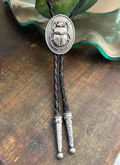 This awesome bolo tie has a meal beetle pendant. Quality guaranteed, picture does not do this buckle justice! The pendant piece measures 1 1/8'' x 1 1/4'' It can adjust lengths, 39'' in total length. The cord is genuine black woven leather.   We have matching belt buckles to pair! Men’s Leather Jewelry, Vintage Black Bolo Ties For Gift, Vintage Black Bolo Ties As Gift, Classic Silver Bolo Tie As Gift, Silver Bolo Ties As A Gift, Brown Bolo Ties As A Gift, Brown Bolo Tie For Gift, Ceramic Bolo Tie, Bolo Tie Outfit