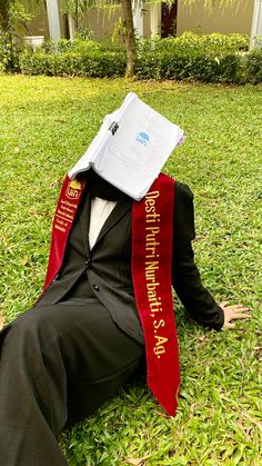 a man sitting in the grass with a book on his head wearing a red sash