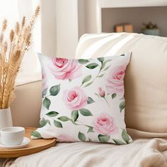 a white pillow with pink roses on it next to a cup and saucer sitting on a couch