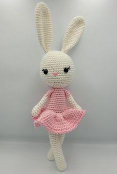 a crocheted bunny doll with a pink dress and big eyes is posed in front of a gray background