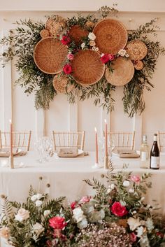 an arrangement of flowers and candles on a table with wicker baskets hanging above it