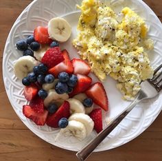 a white plate topped with fruit and scrambled eggs next to a fork on a wooden table