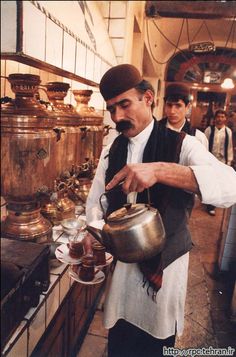 a man in an old - fashioned kitchen pouring tea into a pot with other men looking on