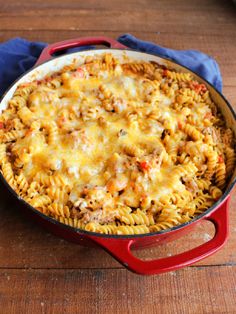 a red skillet filled with pasta and cheese on top of a wooden table next to a blue towel