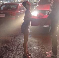 a man and woman standing next to each other in front of a red car at night