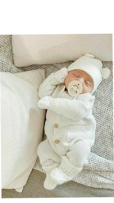 a baby in a white outfit laying on top of a bed next to pillows and blankets