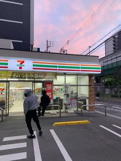 two people are walking in front of a convenience store at dusk with the sun setting behind them