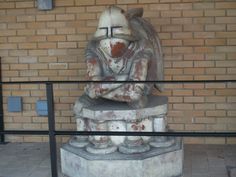 an old statue is sitting in front of a brick wall and metal railings on the sidewalk