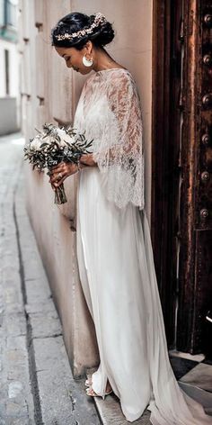 a woman standing next to a building holding a bouquet of flowers and wearing a white dress