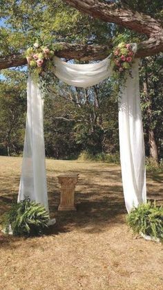 an outdoor wedding setup with white drapes and flowers on the arbor, surrounded by greenery