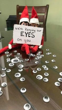 an elf sitting on top of a glass table holding a sign that says, we're keeping our eyes on you