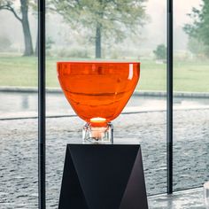 an orange vase sitting on top of a black stand next to a window with trees in the background