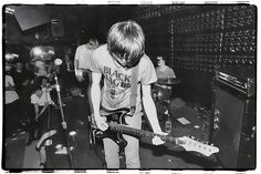 black and white photograph of young man playing guitar in recording studio with other musicians behind him