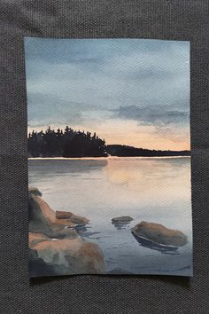 a watercolor painting of a lake with rocks in the foreground