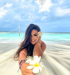 a beautiful woman laying on top of a sandy beach next to the ocean with a flower in her hand